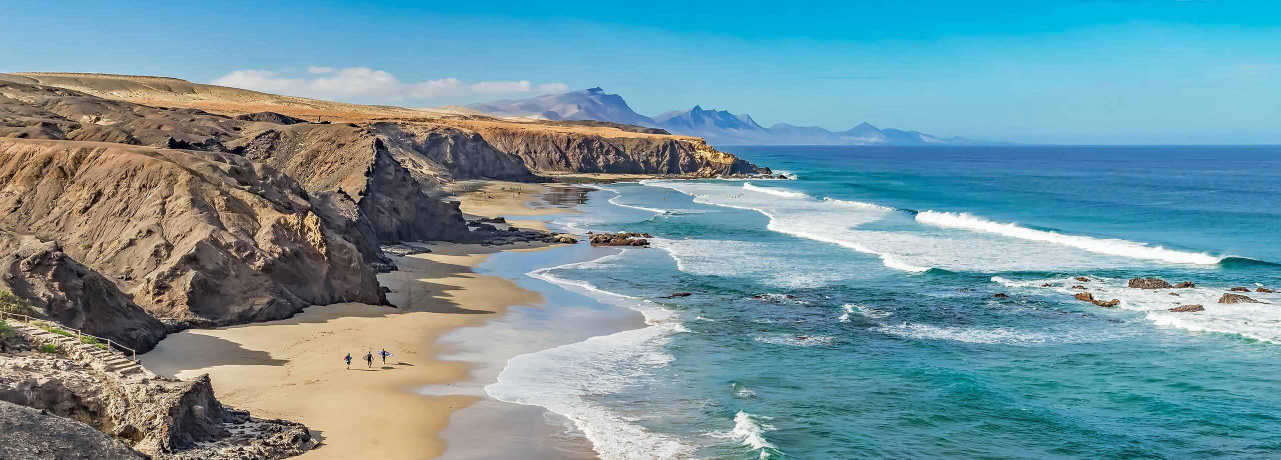 Ein Sandstrand mit auf dem Surfer entlang gehen. Neben dem Strand sind große Felsen zu sehen.