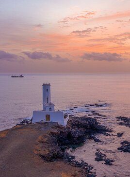 Ein weisser Turm mit einer Mauer drumherum beim Sonnenuntergang. Im Hintergrund ist ein Meer. 