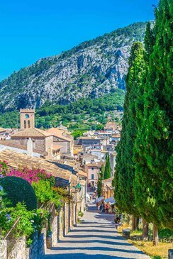 Die Carrer del Calvari Treppe mit Häusern links und Bäumen rechts. Im Hintergrund sind Berge.