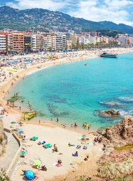 Ein Sandstrand auf der linken Seiten mit Badegästen. Rechts ist ein türkises Meer. Im Hintergrund sind Hotels, Häuser und Berge.