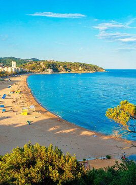 Ein Sandstrand auf der linken Seiten mit Badegästen. Rechts ist ein hellblaues Meer. Im Hintergrund sind Hotels, Häuser und Berge.